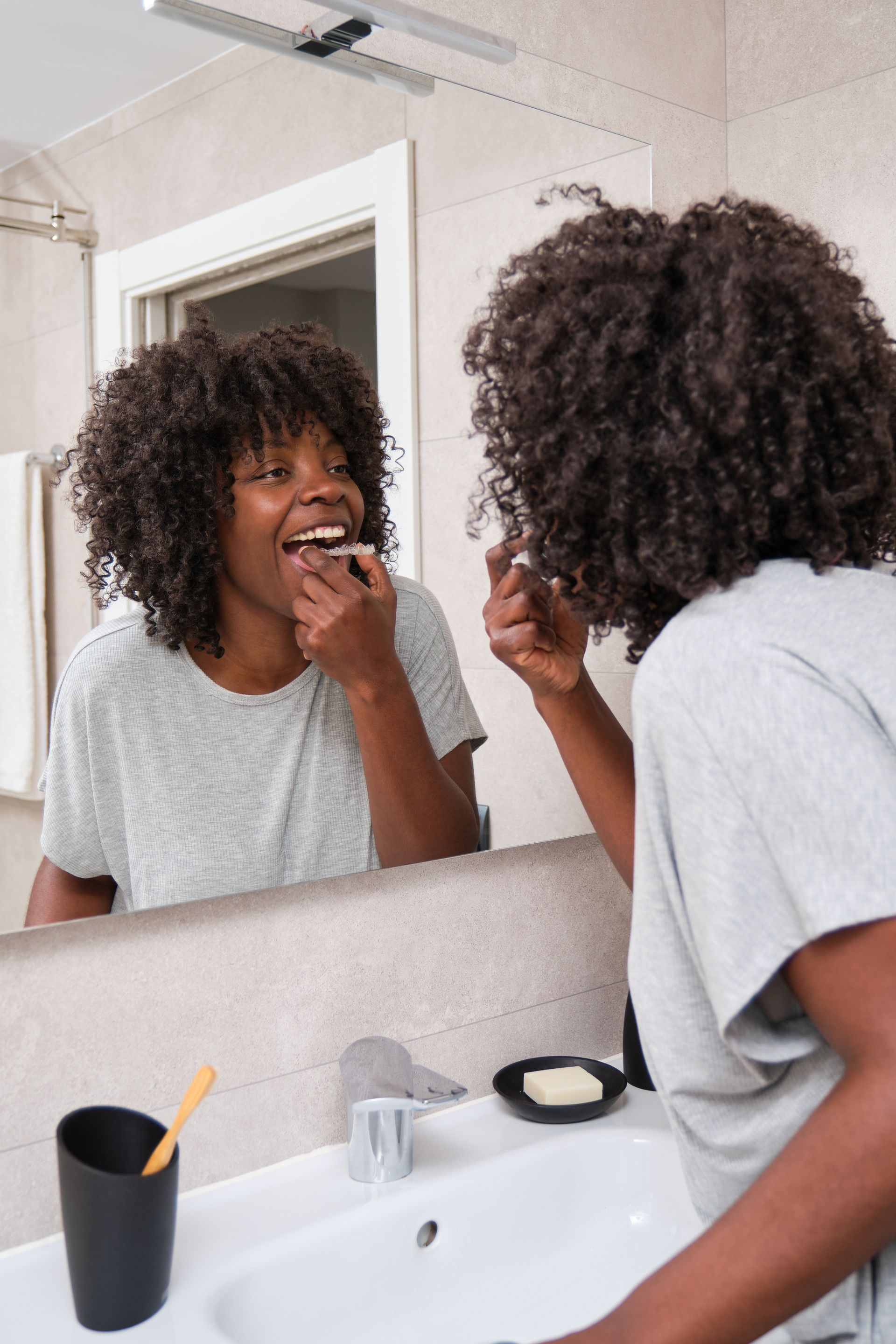 Woman brushing teeth in bathroom, looking in the mirror. She wears a grey t-shirt and smiles.