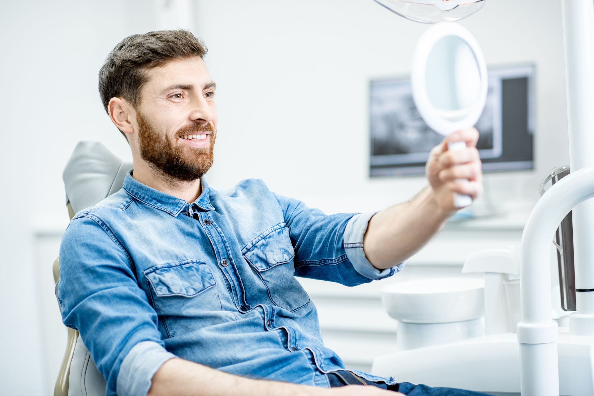 Man in dentist's chair looking at teeth in hand mirror; office setting.