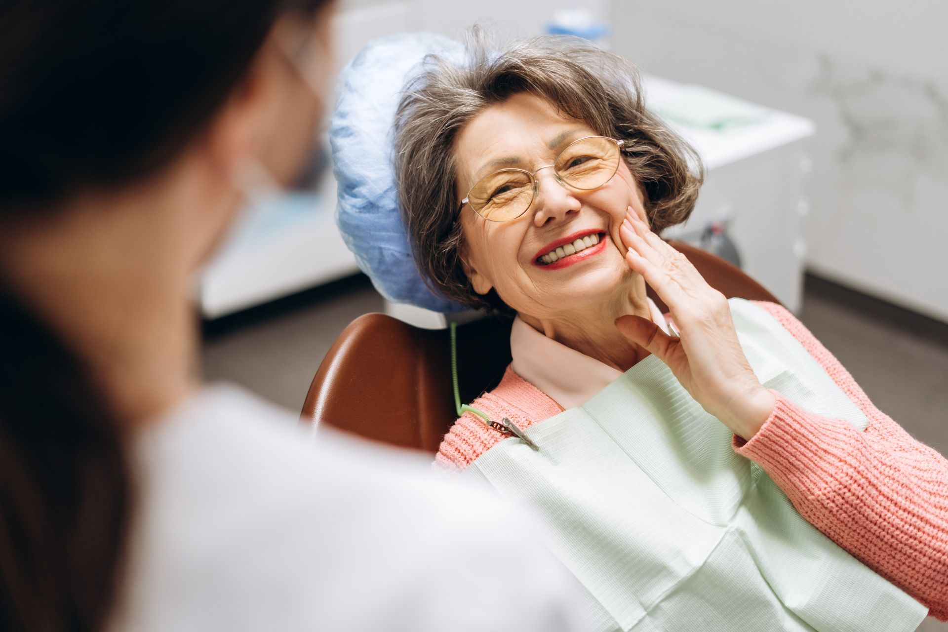 Woman in dentist chair smiles, touching her cheek. Dentist in mask is looking at her.