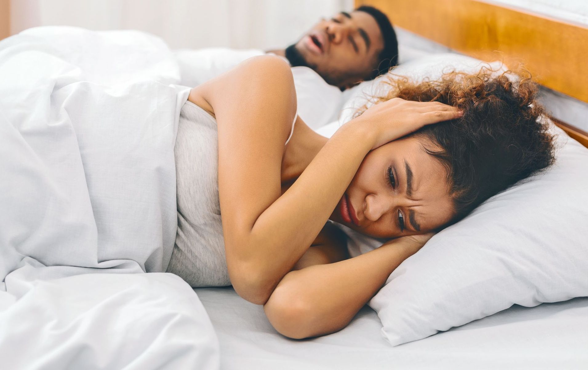 Woman covering ears in bed, man snoring beside her.