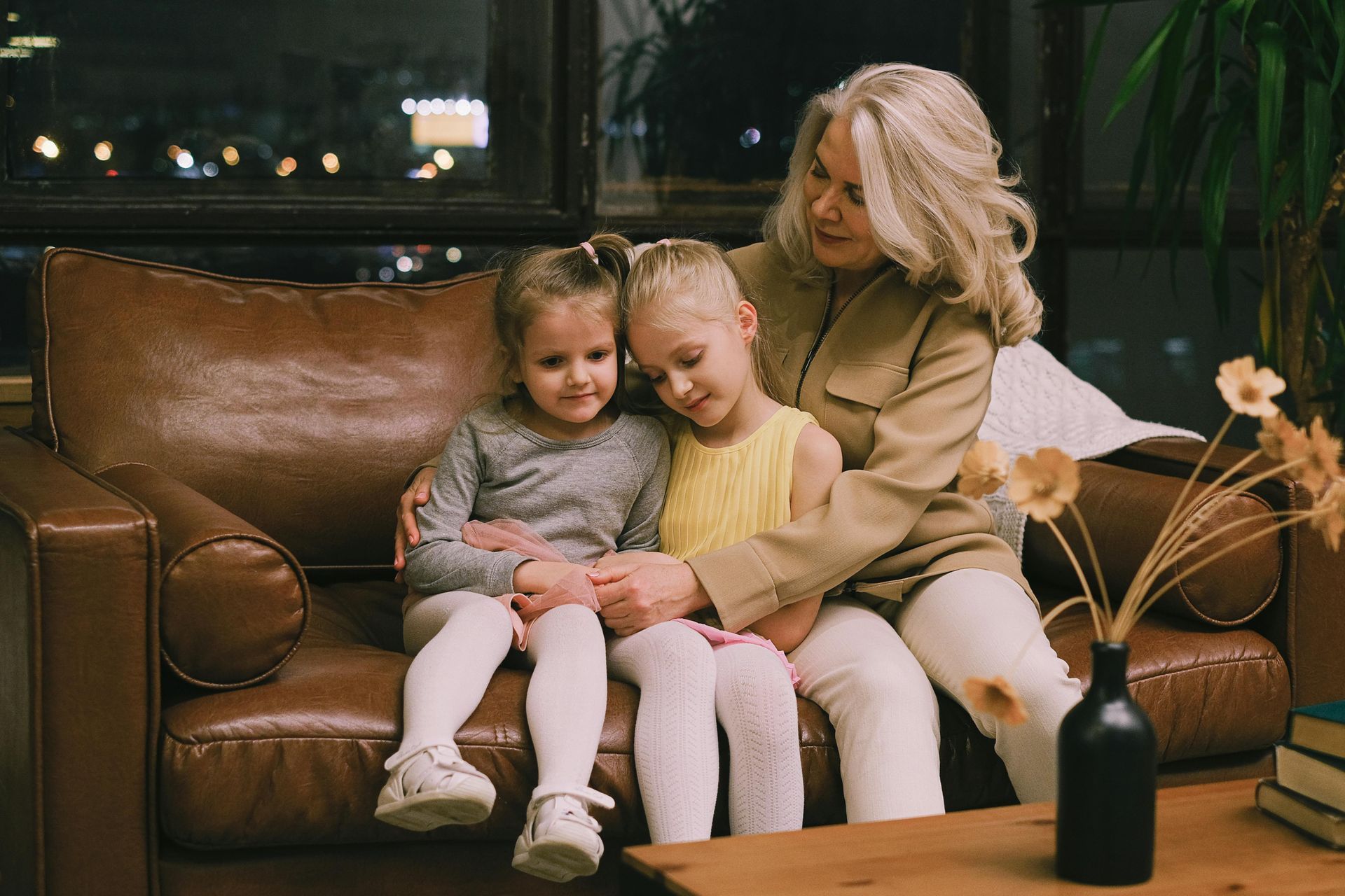 An adult with grey hair sits on a leather couch, embracing two children who are leaning in together.