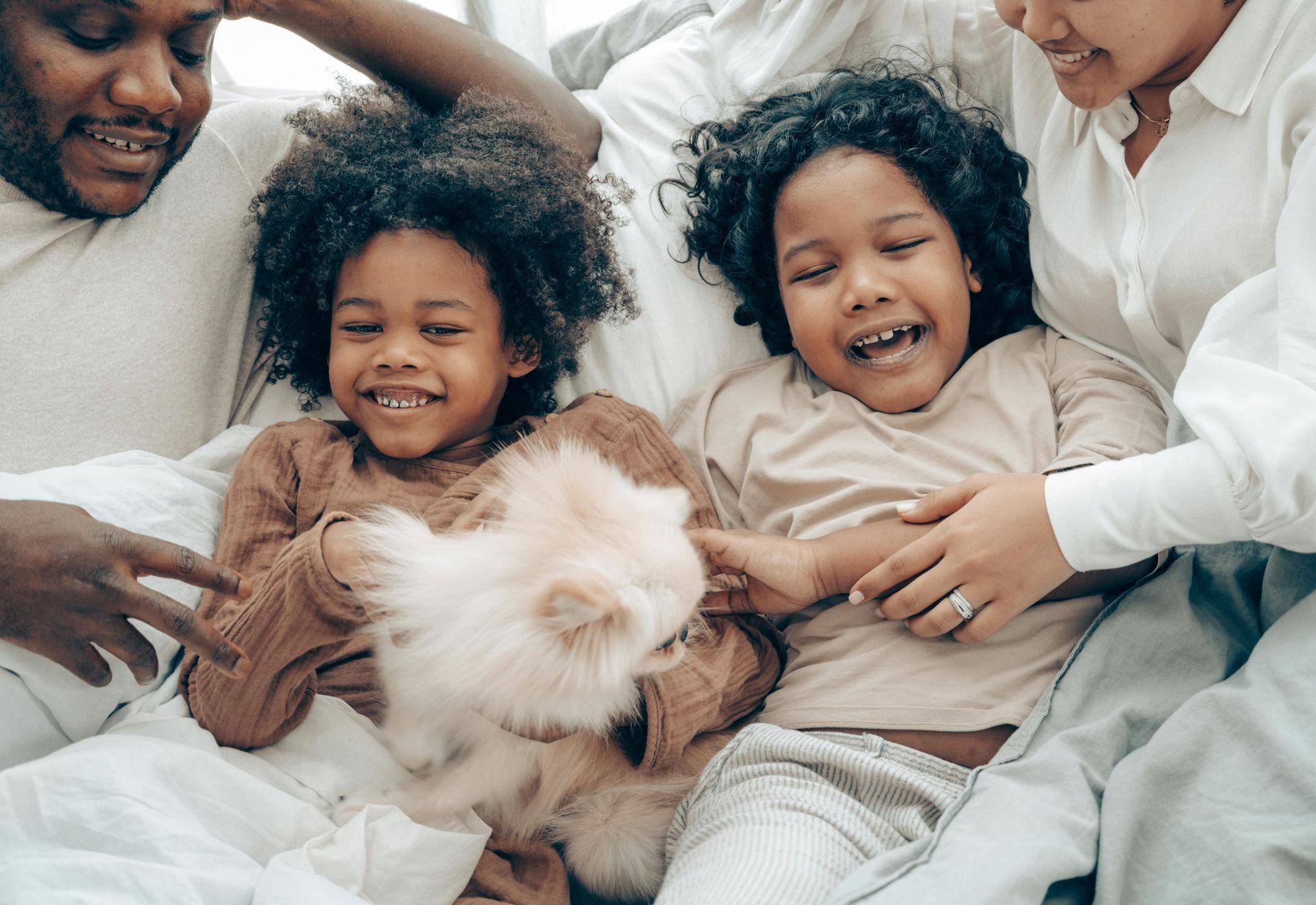 Two children laugh while holding a small, fluffy white dog on a bed with their parents nearby.