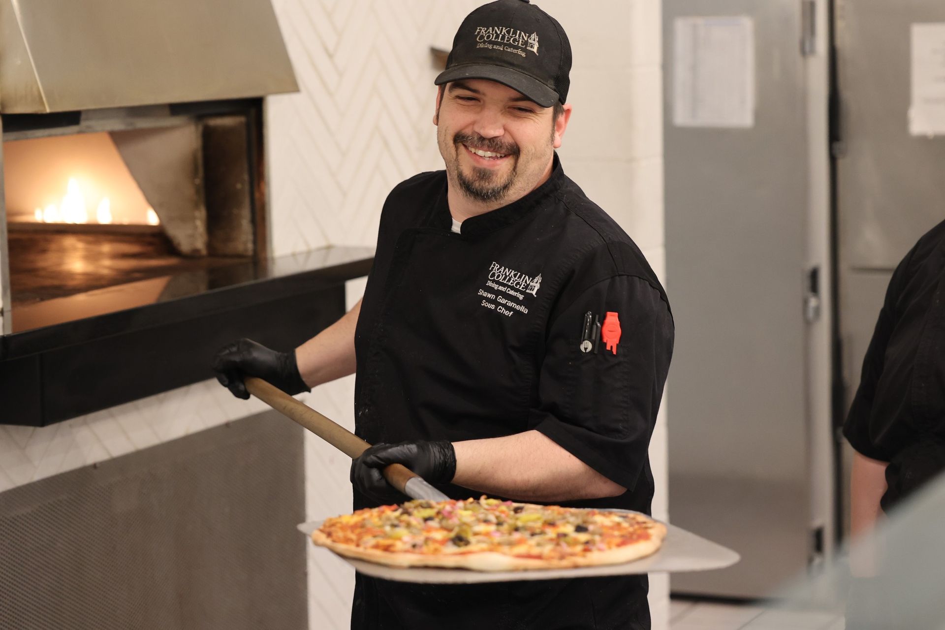 Chef smiles while holding a pizza on a peel near a pizza oven. He's wearing a black uniform and hat.