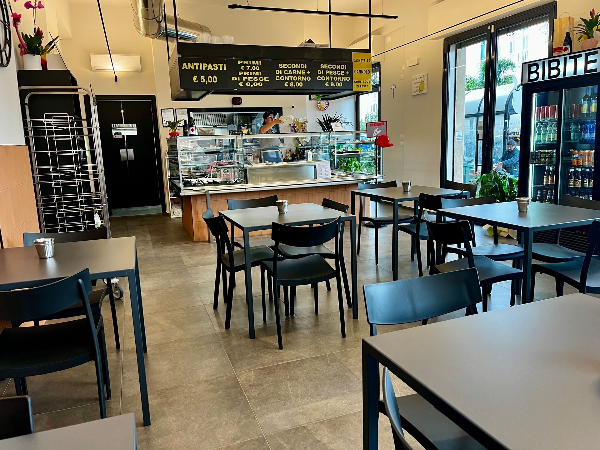 Interno di un bar con tavoli e sedie. Sullo sfondo il bancone con l'esposizione dei prodotti alimentari. Finestra sulla destra.