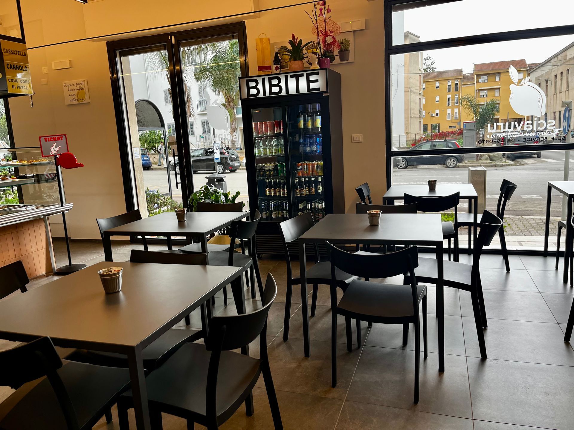 Interno di un bar con tavoli, sedie e un refrigeratore per bevande, con vista sulla strada esterna.