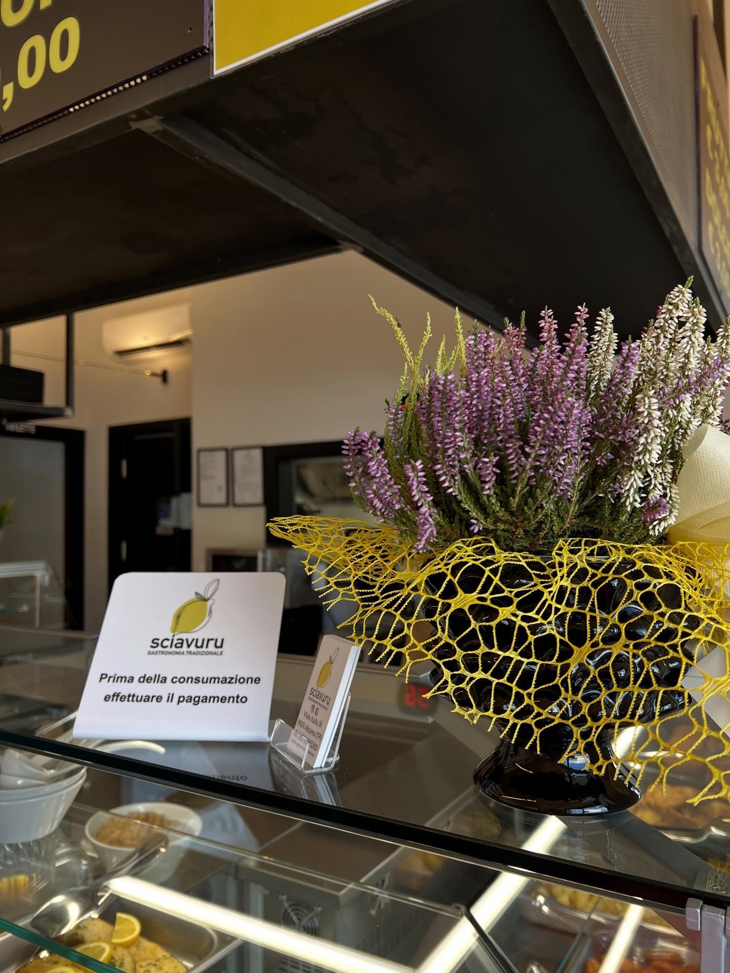 Un vaso di fiori viola e una rete gialla sul bancone di un negozio. È esposto un cartello con il logo di un limone.