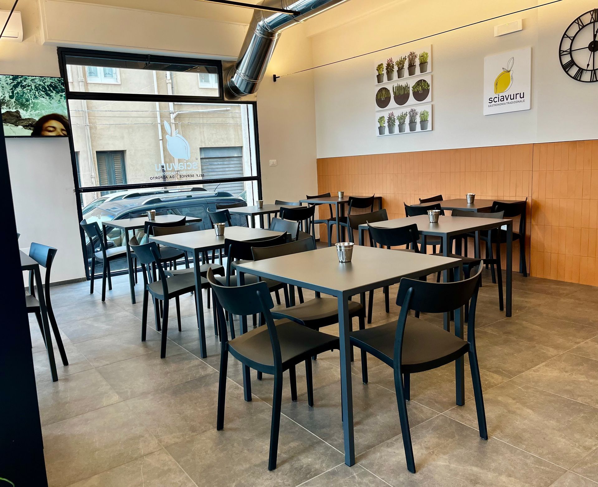 Interno del ristorante: tavoli, sedie e ampia finestra. Pareti color crema e arancione. Sono presenti un orologio e opere d'arte.