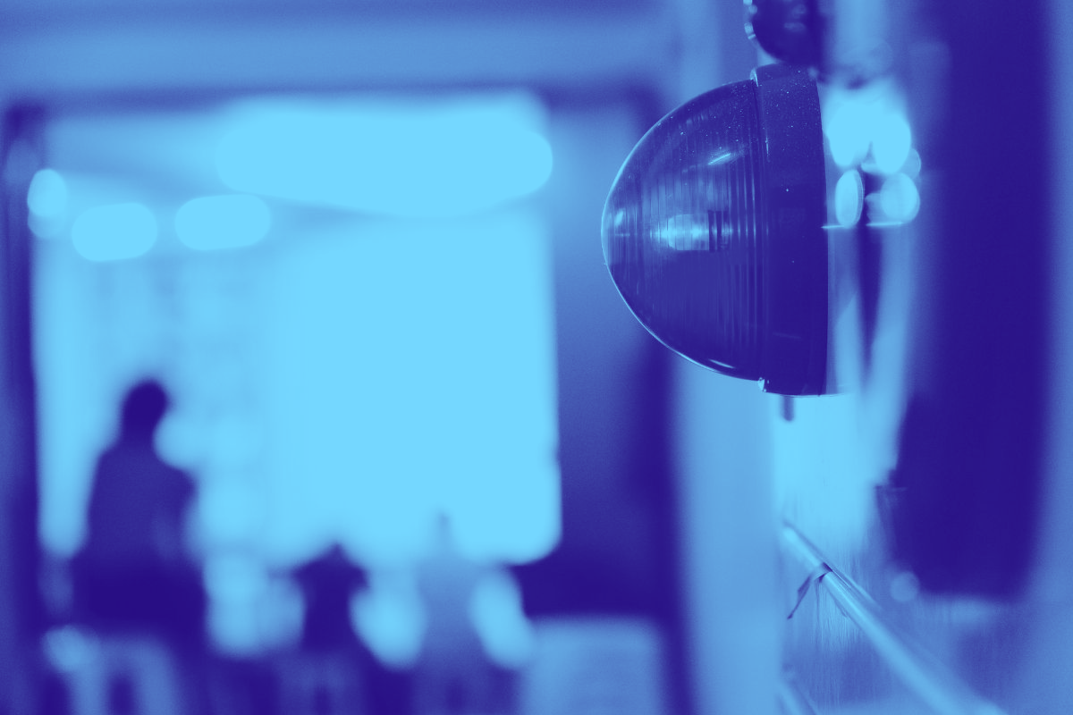 Blue-toned view of an industrial light fixture with people blurred in the background, possibly indoors.