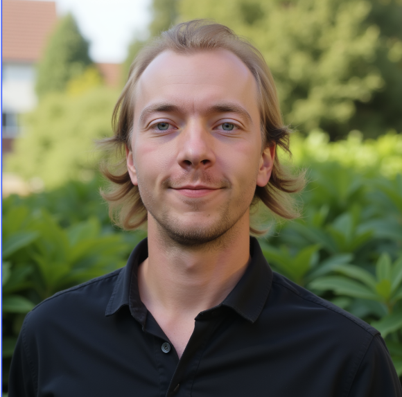 Man with blond hair, wearing a black shirt, smiling in front of green foliage.