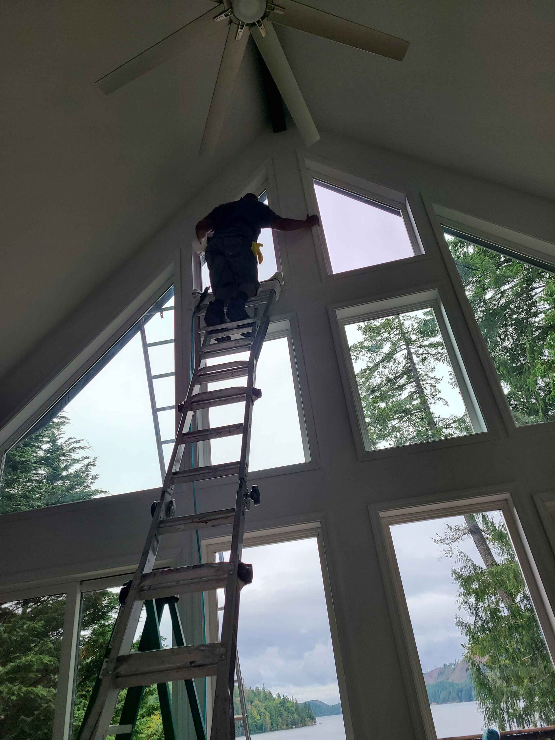 Person on ladder installing window tint in a room with tall, multi-paned windows overlooking a lake.