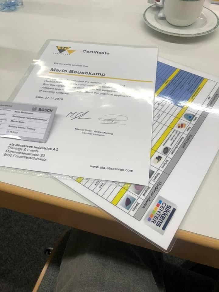 Een certificaat voor Mario Braukamp, ​​een klein naamplaatje en een overzichtstabel liggen op een witte tafel naast een kopje thee.