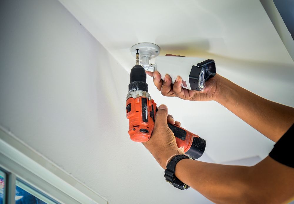 Hands Using a Drill to Mount a White Smoke Detector to A Ceiling — AC/DC Electrical, Air Conditioning & Refrigeration In Esk, QLD