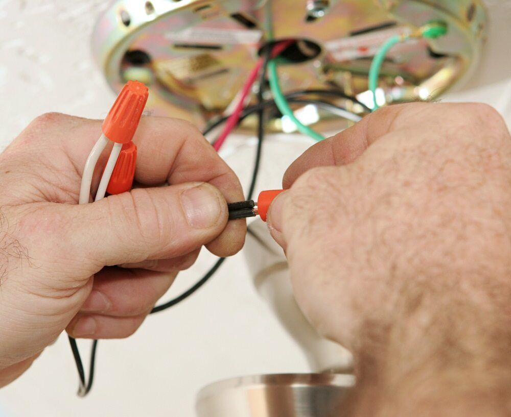 Close-Up of Hands Connecting Electrical Wires with An Orange Wire Nut on A Ceiling Light Fixture — AC/DC Electrical, Air Conditioning & Refrigeration In Esk, QLD