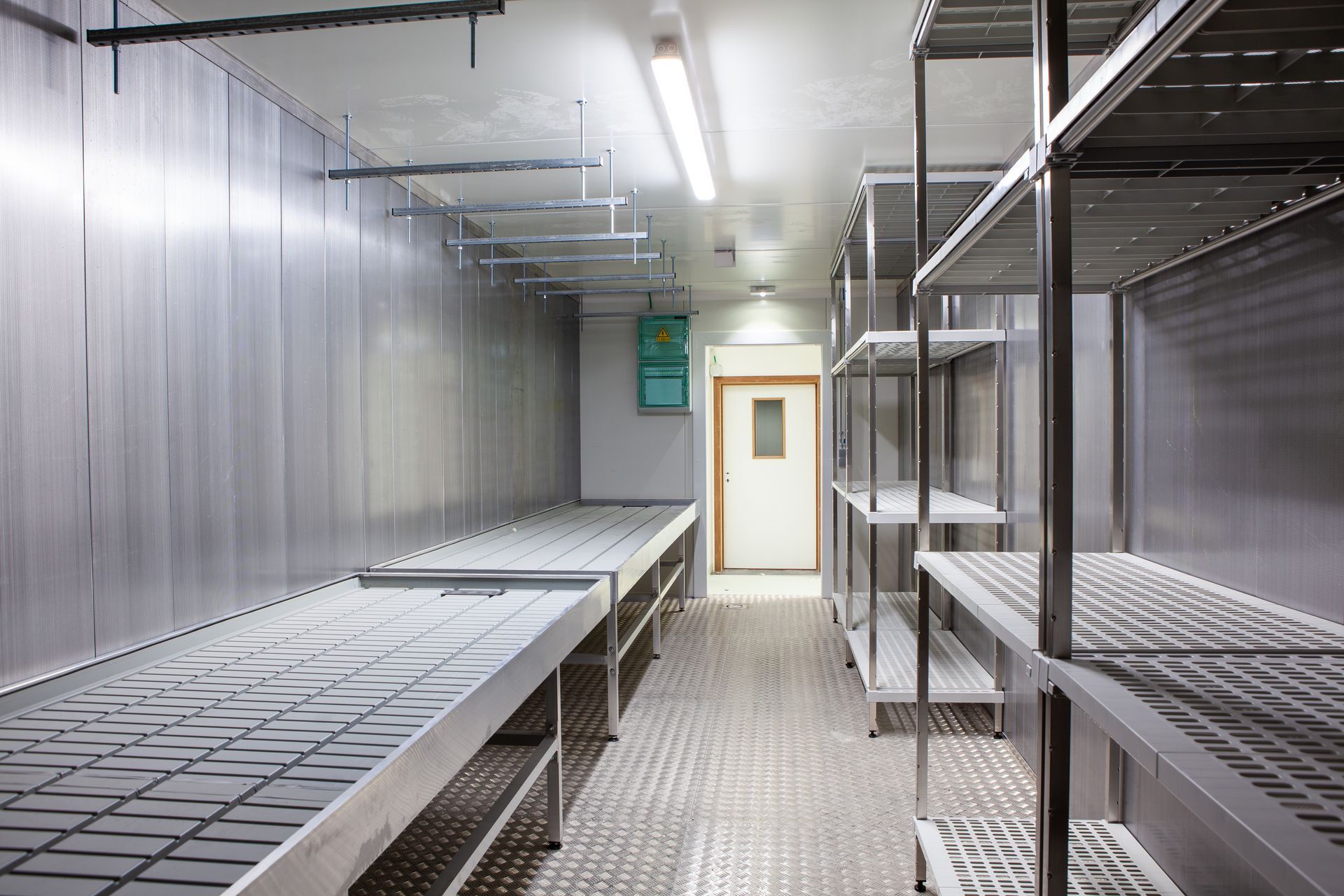 Empty stainless-steel industrial kitchen with metal worktables and shelves, lit by fluorescent lights.