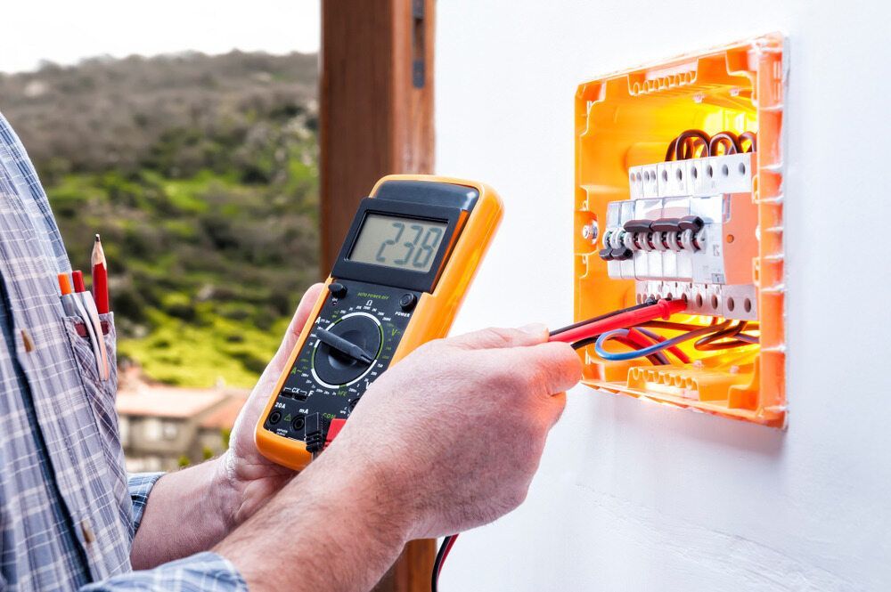 An Electrician Uses a Digital Multimeter to Test the Voltage on A Circuit Breaker Box Mounted on A White Wall — AC/DC Electrical, Air Conditioning & Refrigeration In Toowoomba, QLD