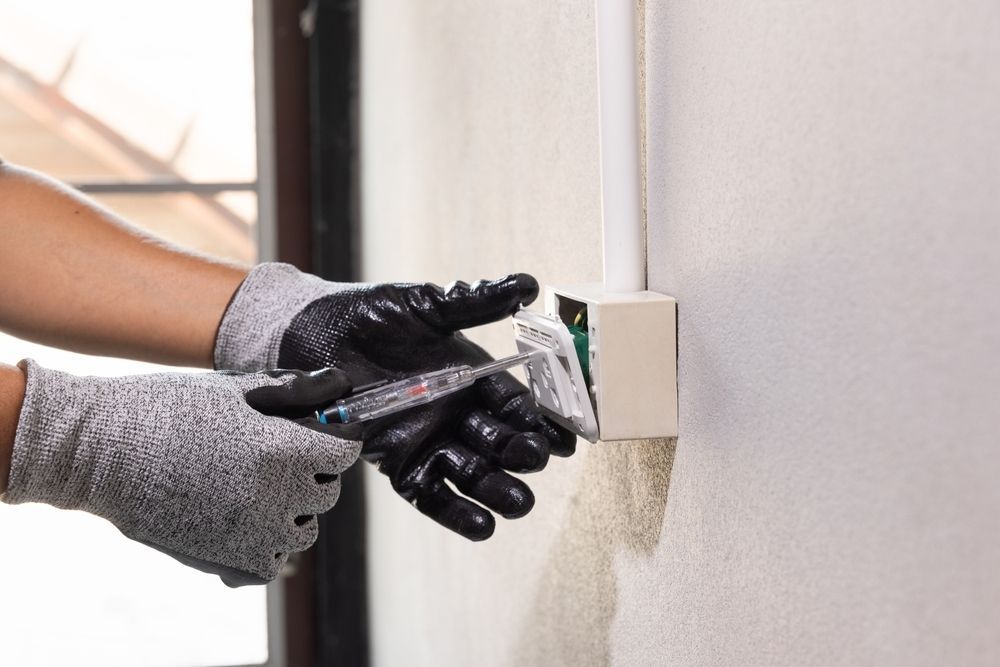 Hands Wearing Protective Work Gloves Using a Voltage Tester on An Electrical Wall Outlet — AC/DC Electrical, Air Conditioning & Refrigeration In Toowoomba, QLD