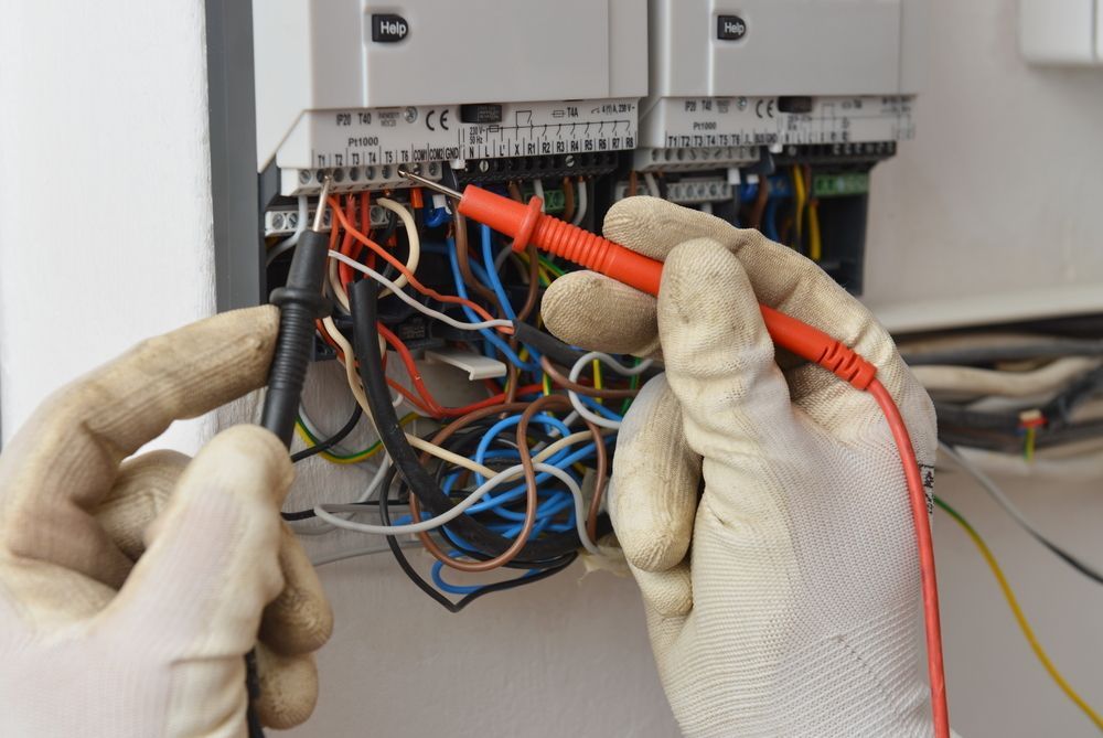 Hands Wearing Work Gloves Test Electrical Connections on A Control Unit with A Multimeter — AC/DC Electrical, Air Conditioning & Refrigeration In Zilzie, QLD
