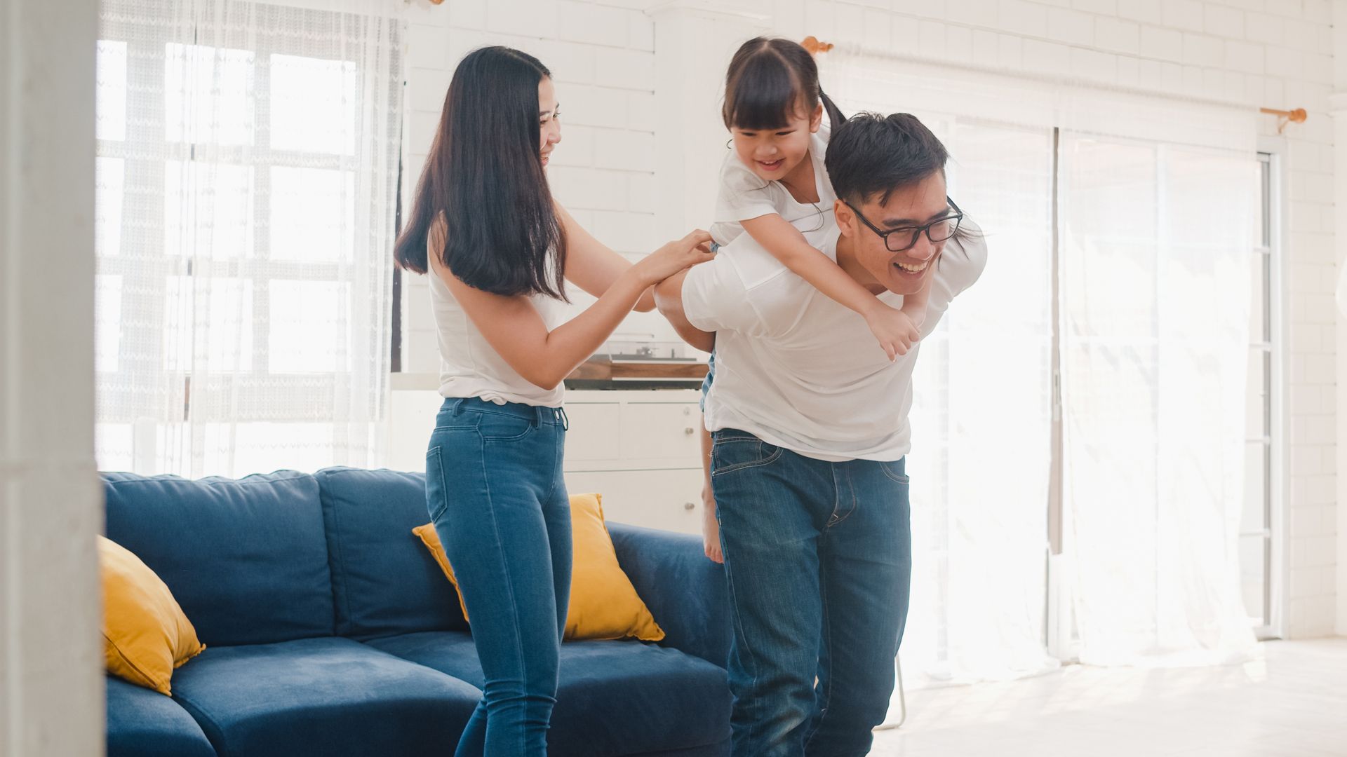 Family playing in a brightly lit living room. Father carries child on his shoulders while mother holds his arm.