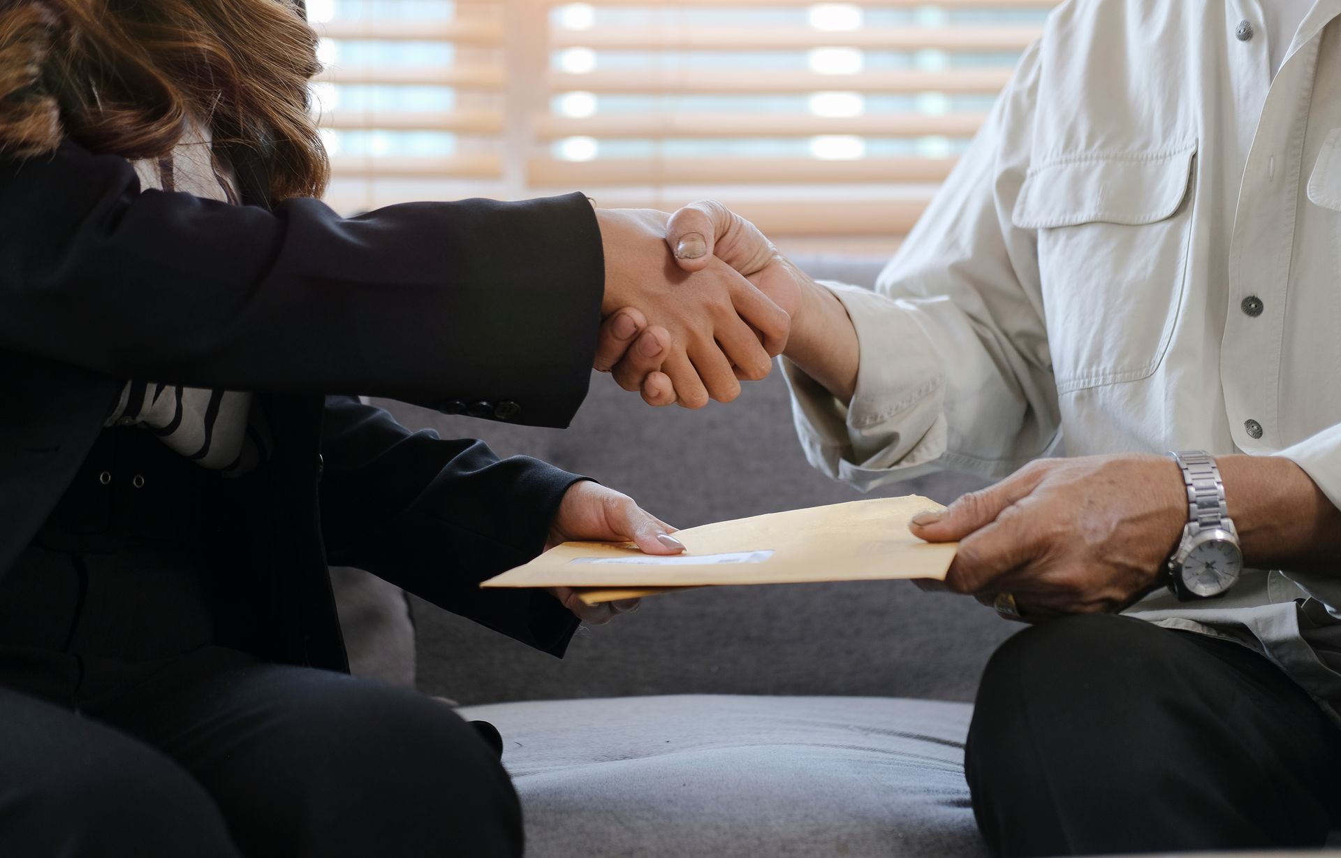 Woman in a black suit shakes hands with a person in a light shirt, handing them an envelope.