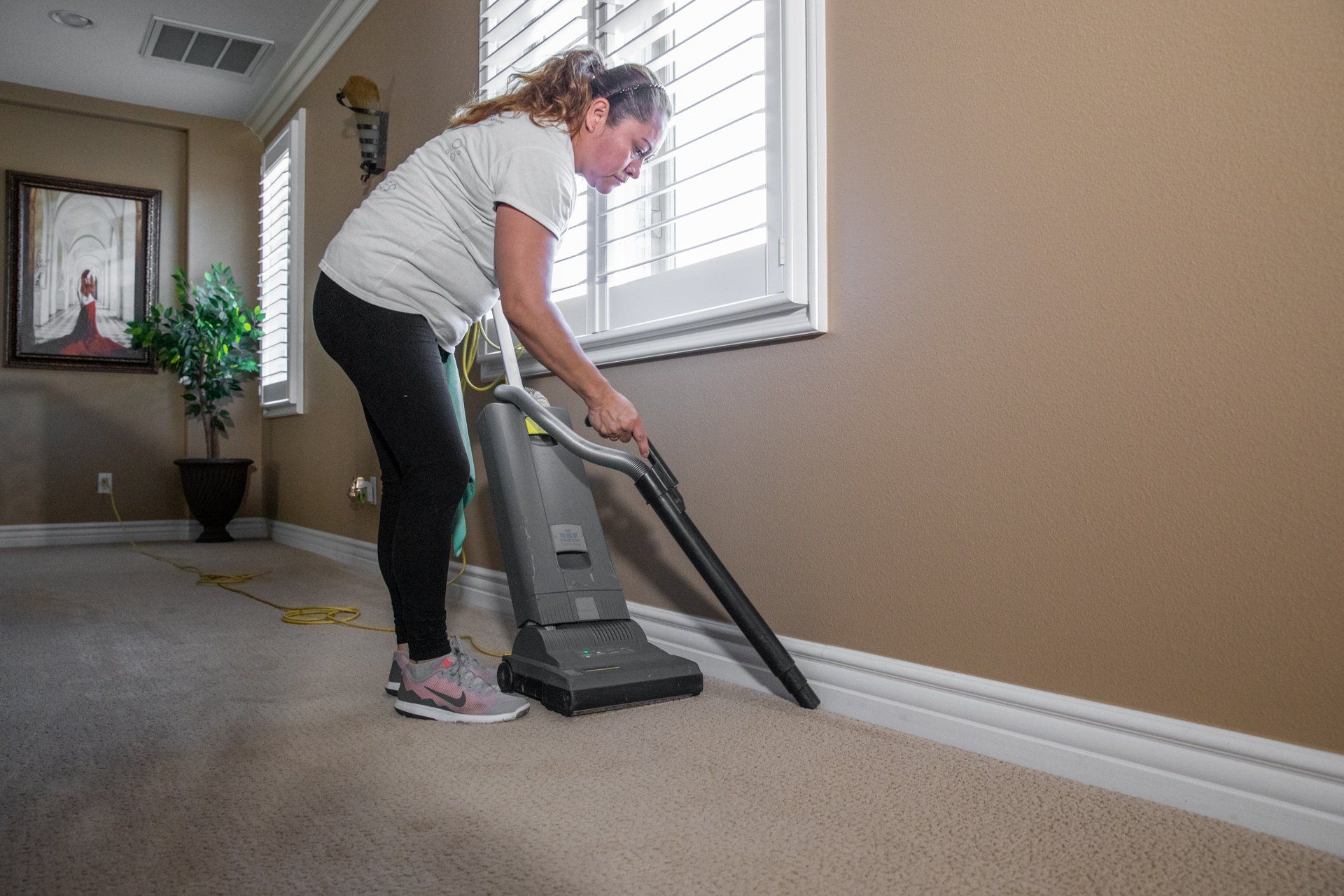 Woman vacuums a beige carpet next to a white wall and window.