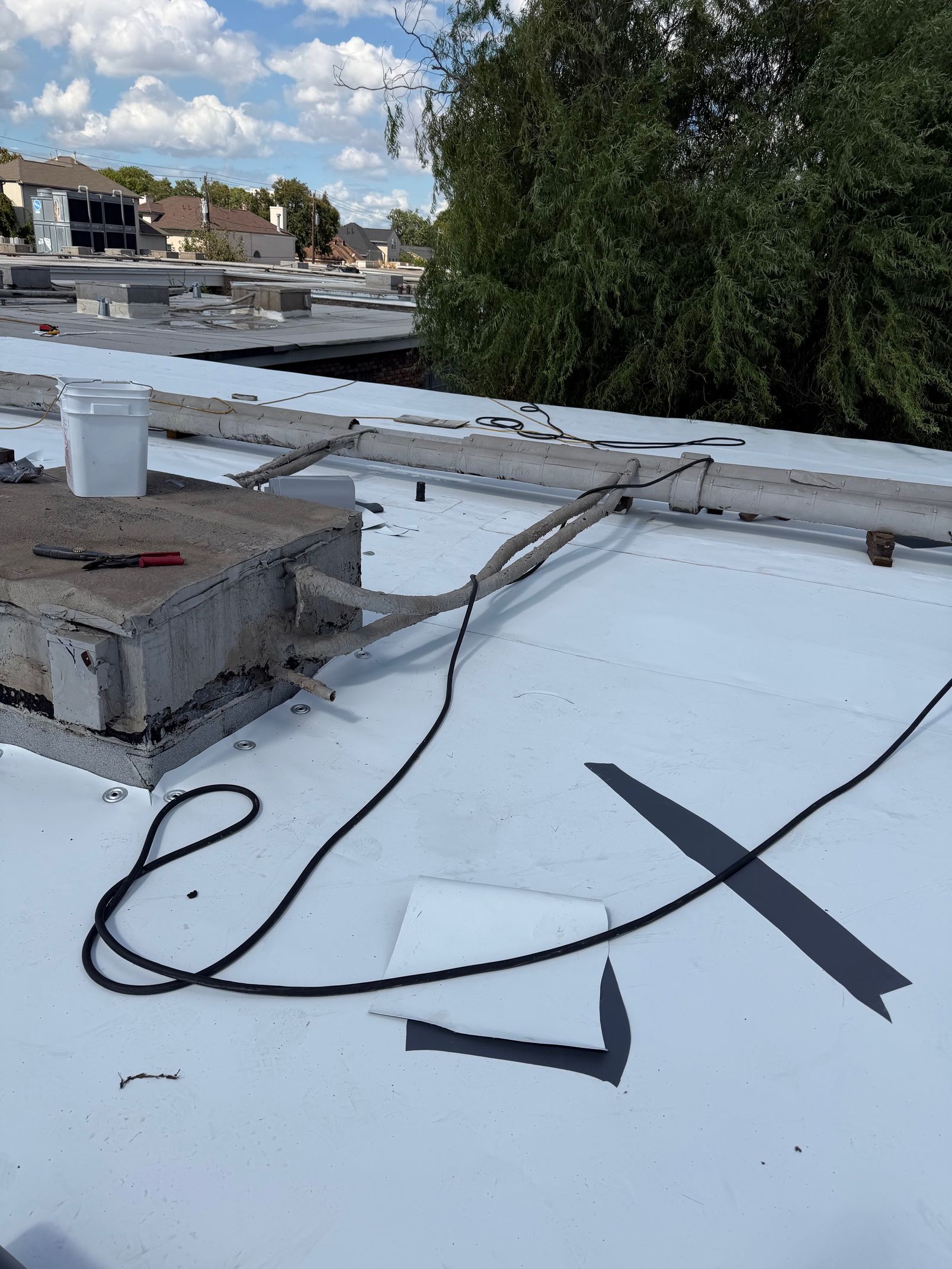 An elevated view of a flat, white rooftop with a concrete structure, exposed cables, and a bucket under a cloudy sky.