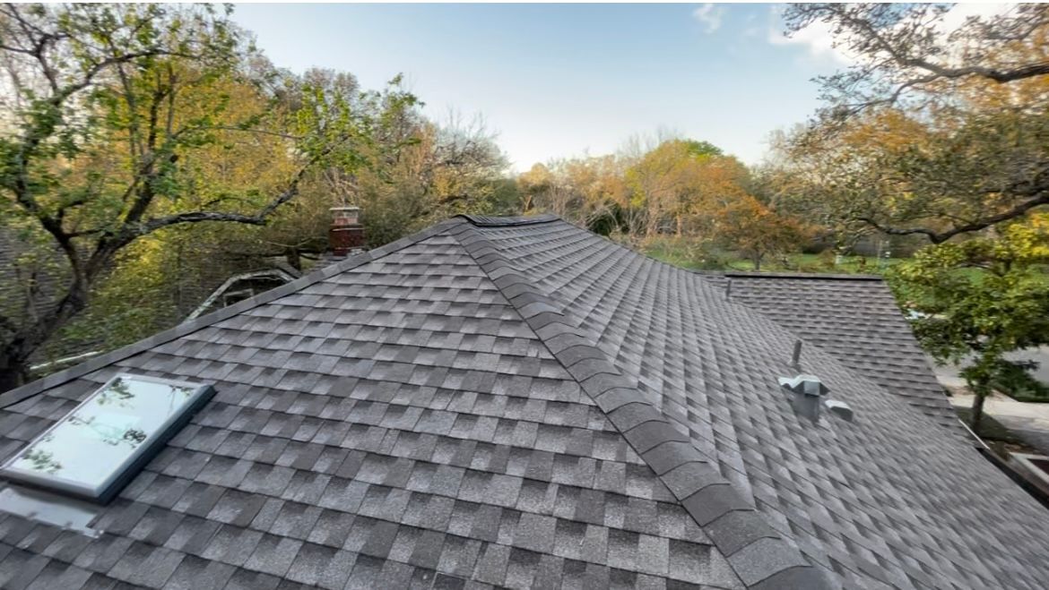 A high-angle view of a gray shingled roof with a skylight, a chimney, and a vent pipe, surrounded by trees.