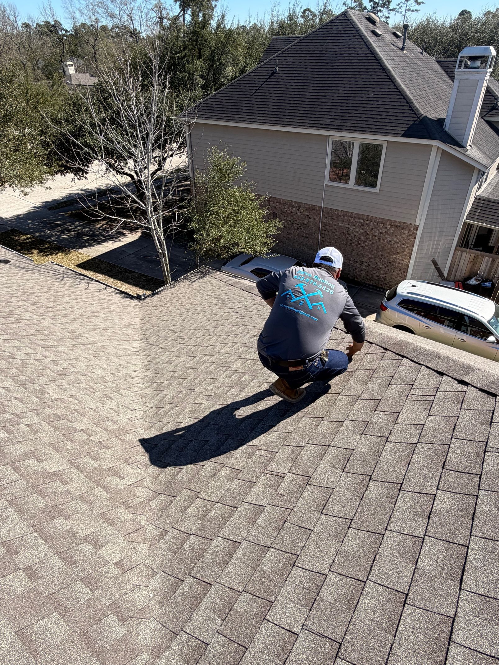 A person wearing a grey shirt and white hat crouches on a textured, shingled residential roof.