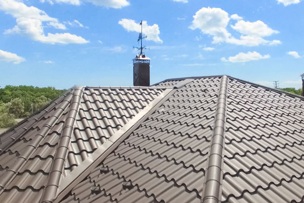 A metal tiled roof under a blue sky, featuring a chimney with an attached weather vane.