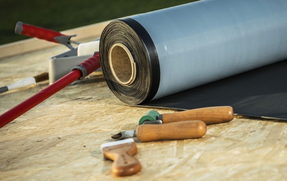 A roll of black roofing membrane lies on a plywood surface with various installation tools, including rollers and brushes.