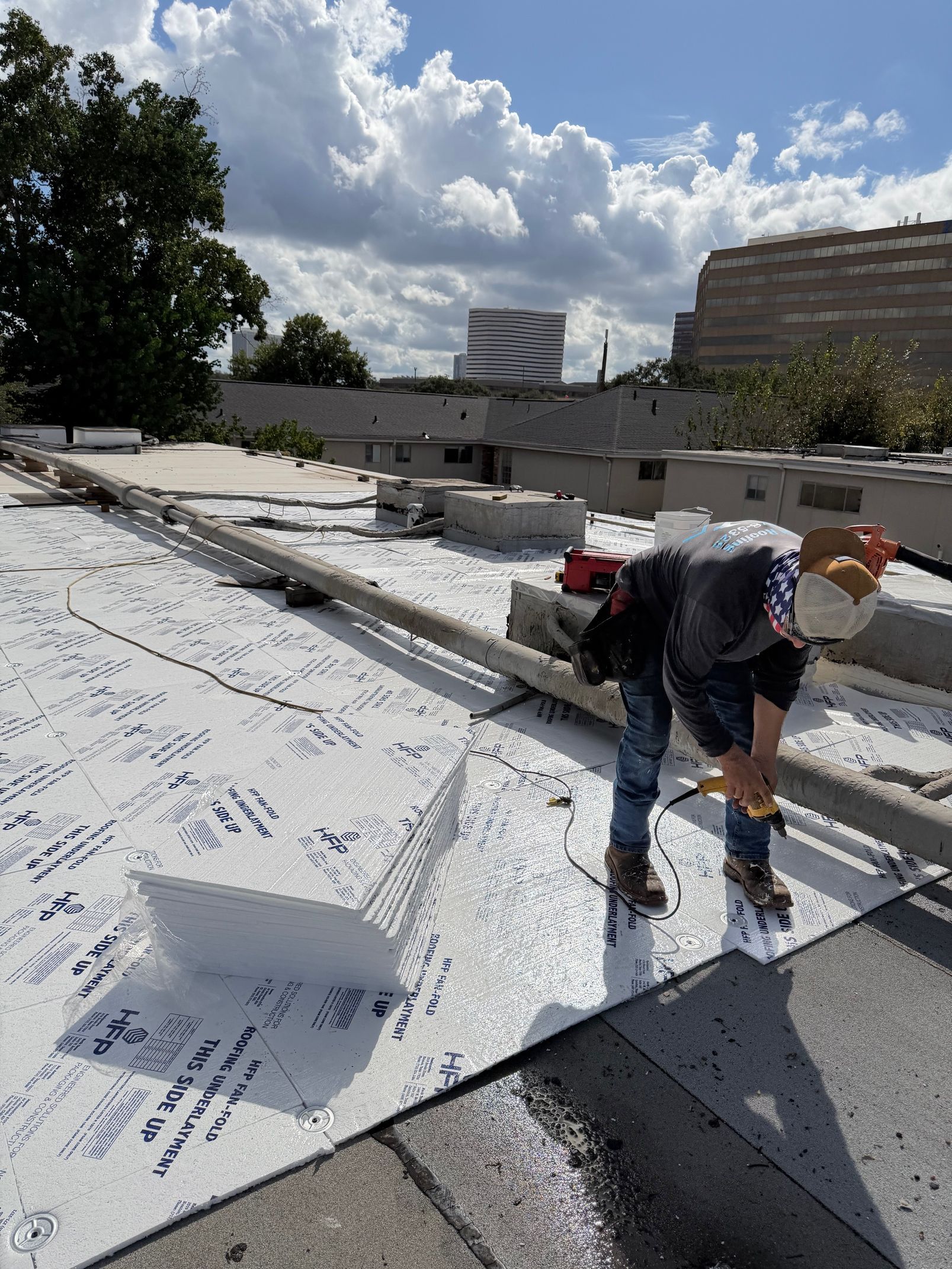 A worker installs white roofing material on a sunny rooftop, with urban buildings and trees in the background.