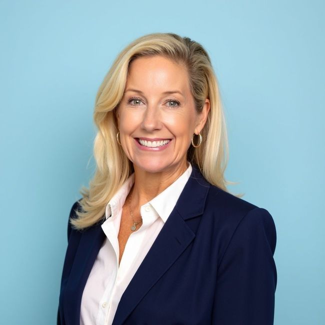 Woman with blonde hair, smiling, wearing a navy blazer and white shirt against a blue background.