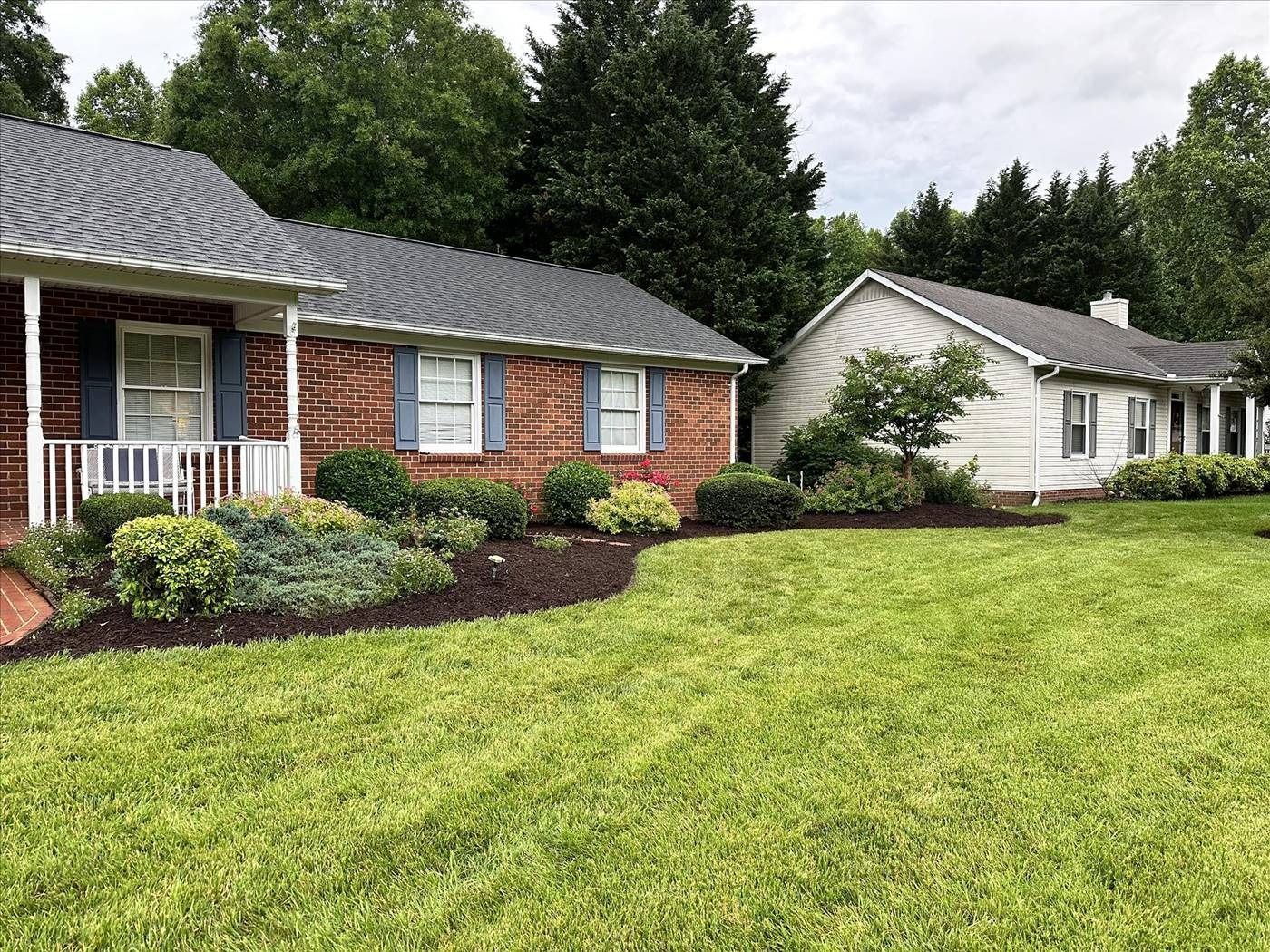 A brick house with a porch and a white house with a lush green lawn in front of it.