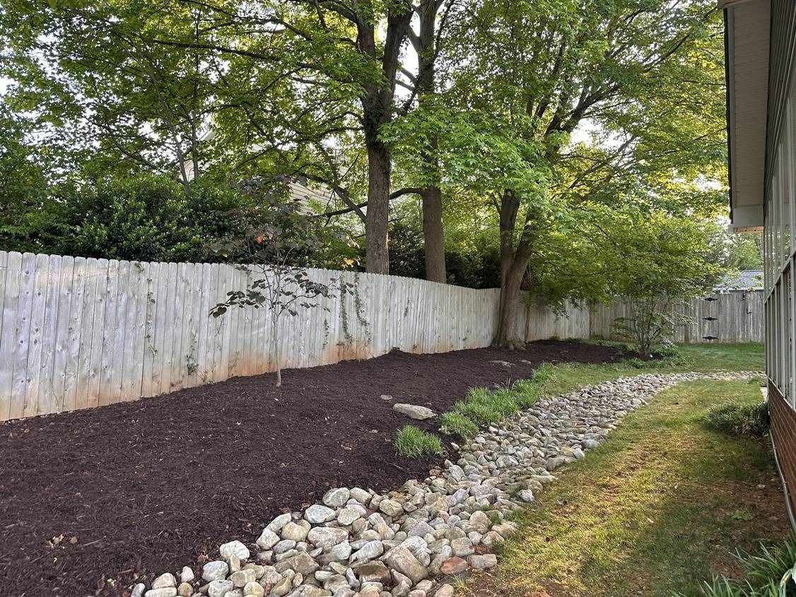 A backyard with a white fence , trees and rocks.