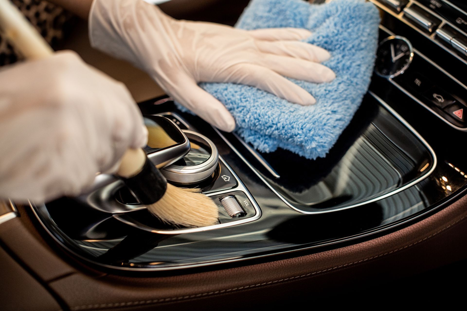 Gloved hands cleaning a car’s center console with a brush and blue microfiber cloth
