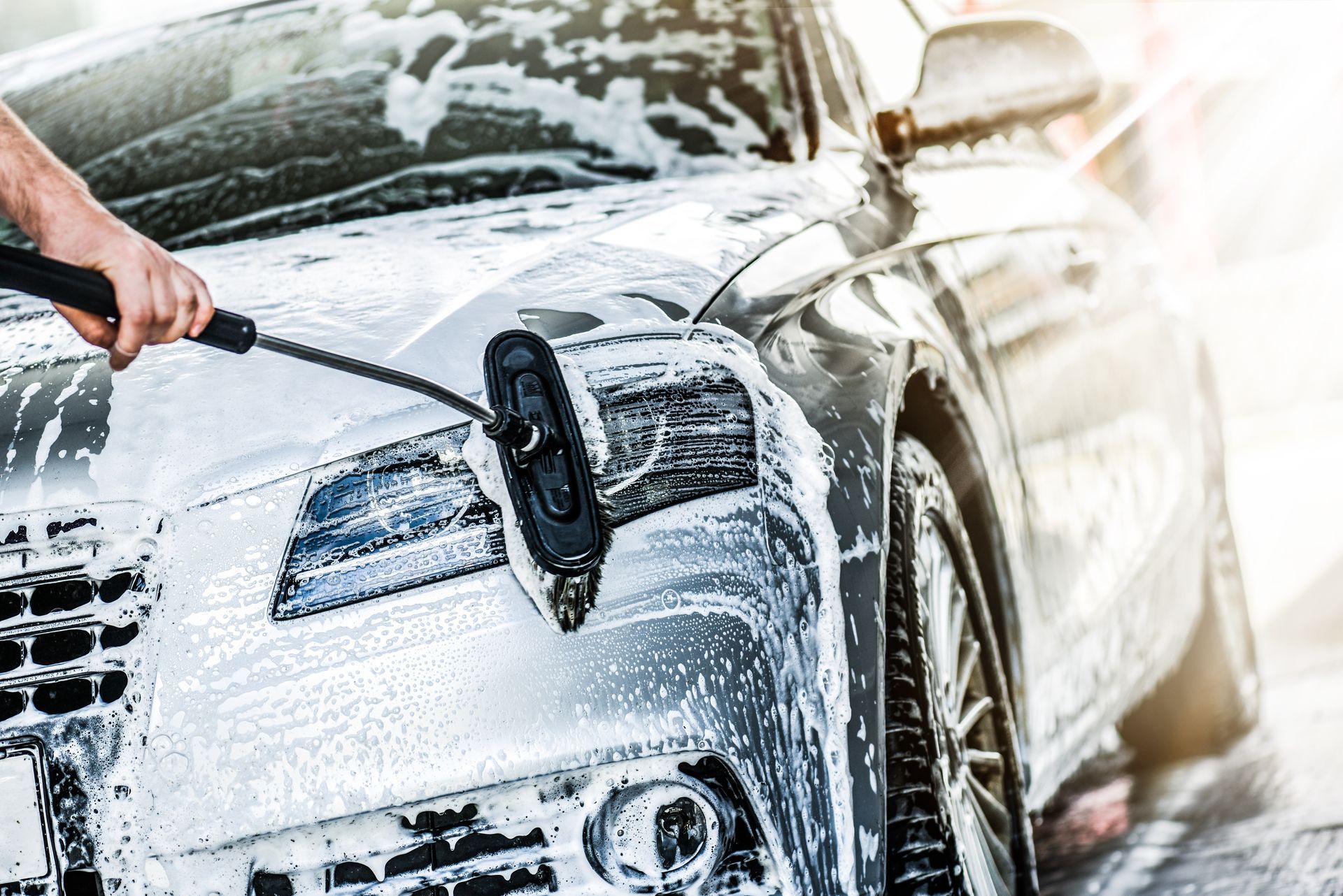 Car being washed with a pressure hose, covered in thick white foam at a car wash.