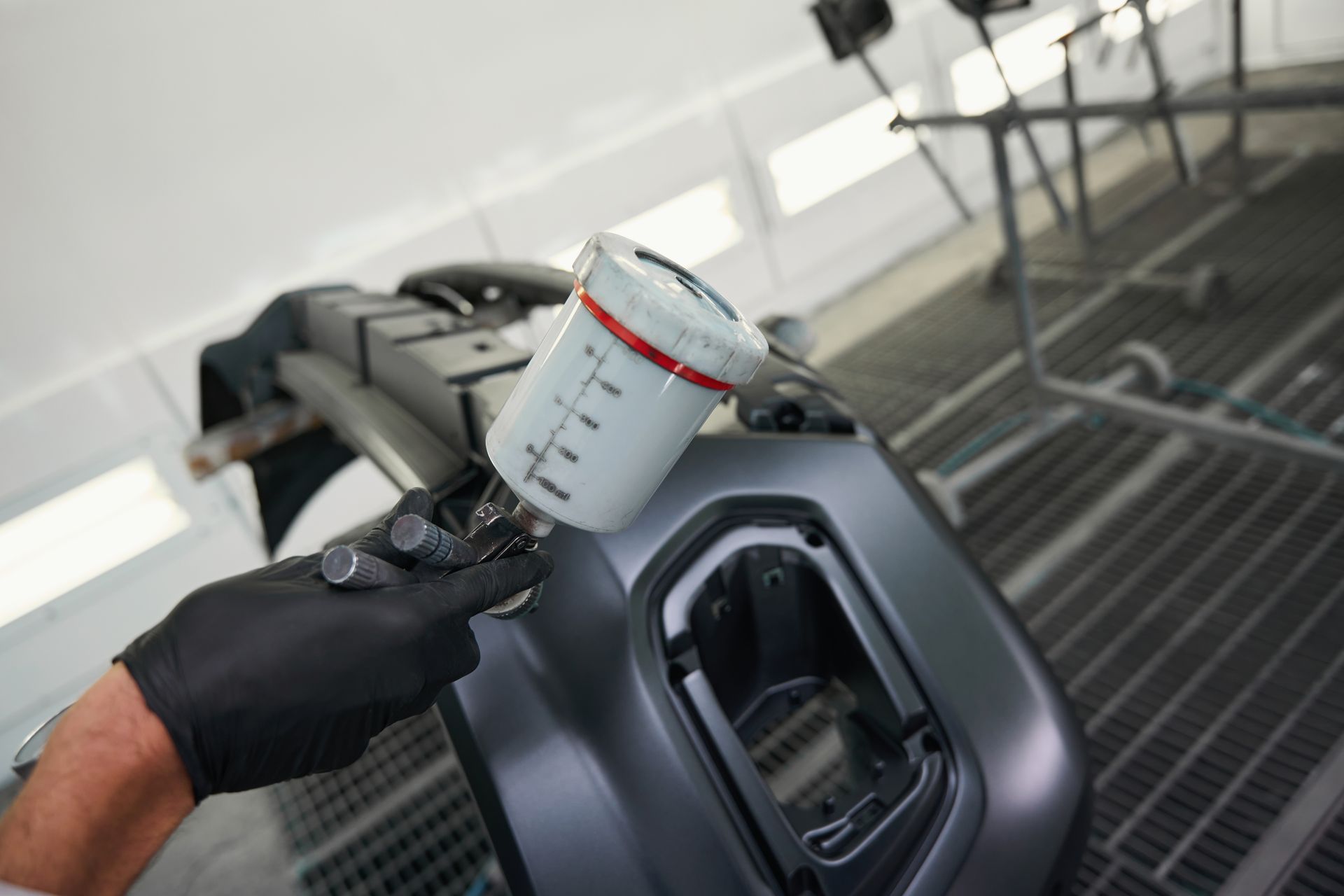 Gloved hand holding a spray gun over a car part in a paint booth