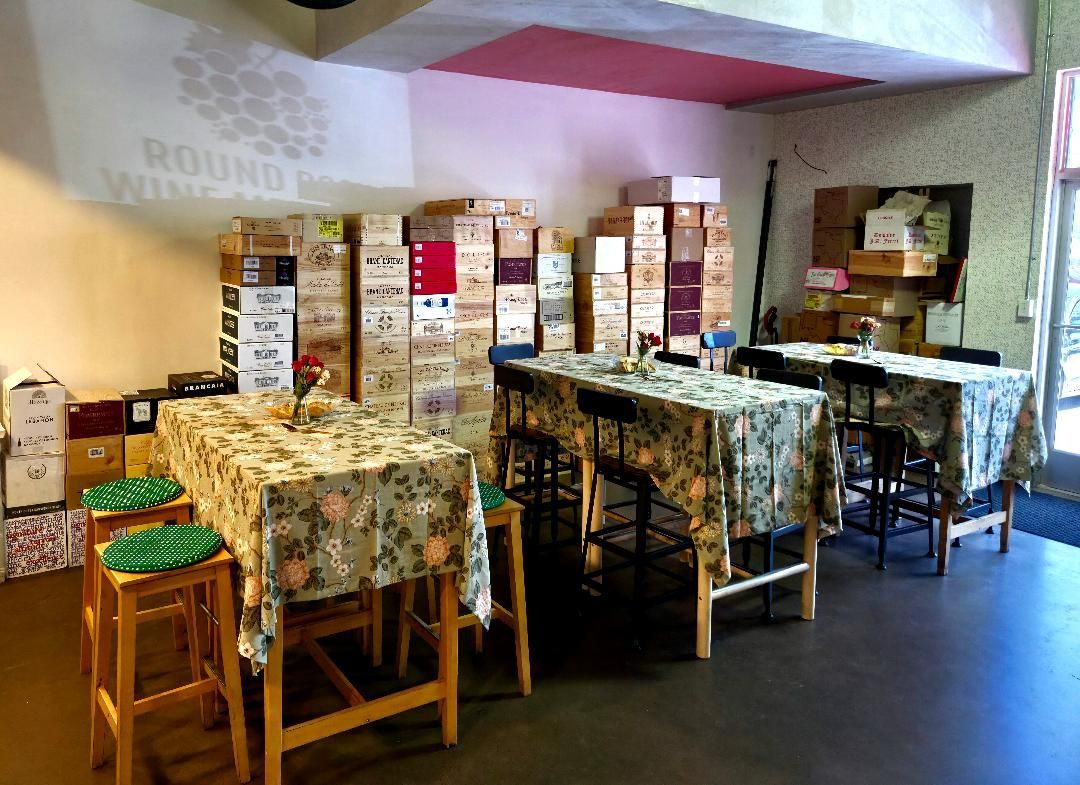 Tables and stools in a wine shop with boxes of wine in the background.