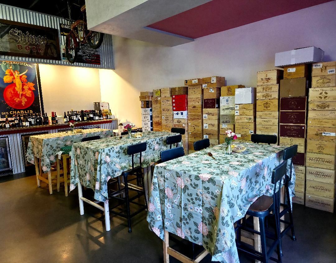 Interior of a wine shop. Tables with floral tablecloths are set up. Boxes of wine are stacked against a wall.