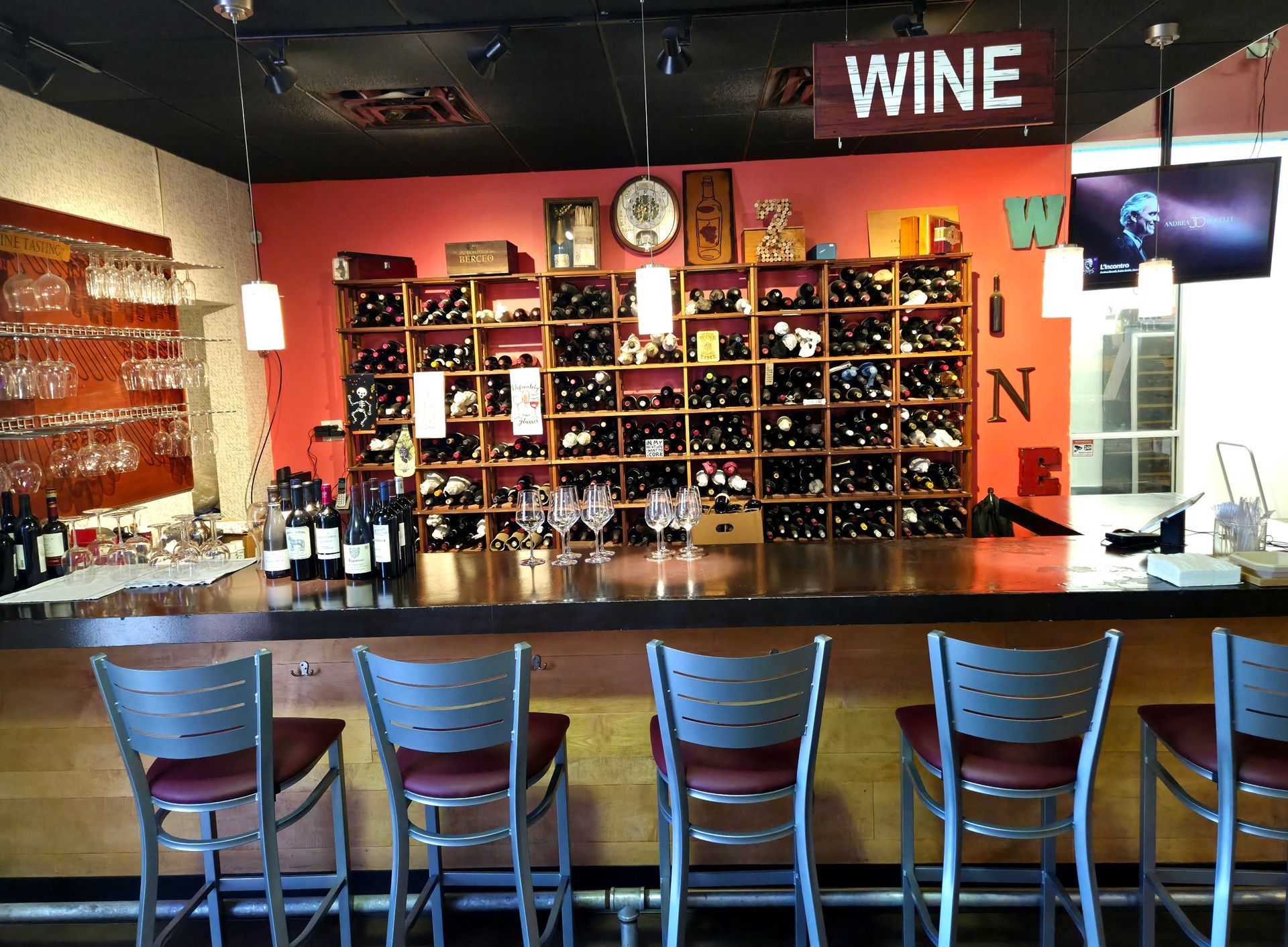 Wine bar interior with bottles on shelves, bar with stools, red wall, and a wine sign.