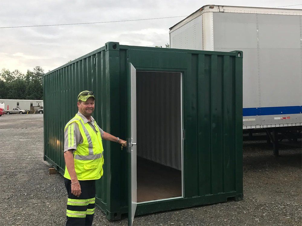 A man in a yellow vest is opening the door of a green shipping container.