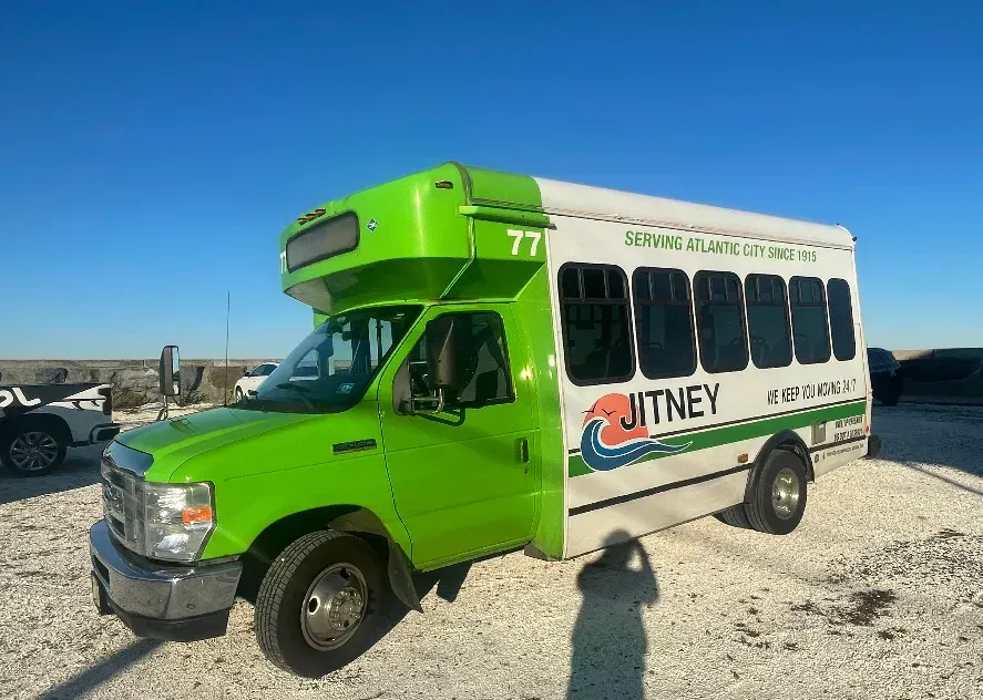 A green and white jitney bus is parked in a gravel lot