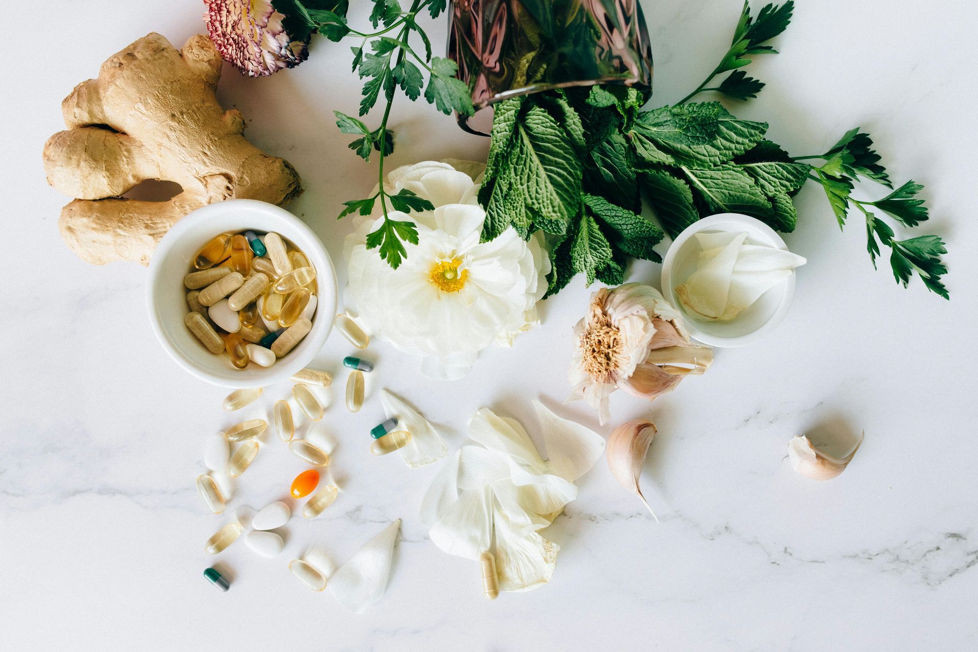 Ginger, herbs, flowers, and supplements arranged on a white marble surface.
