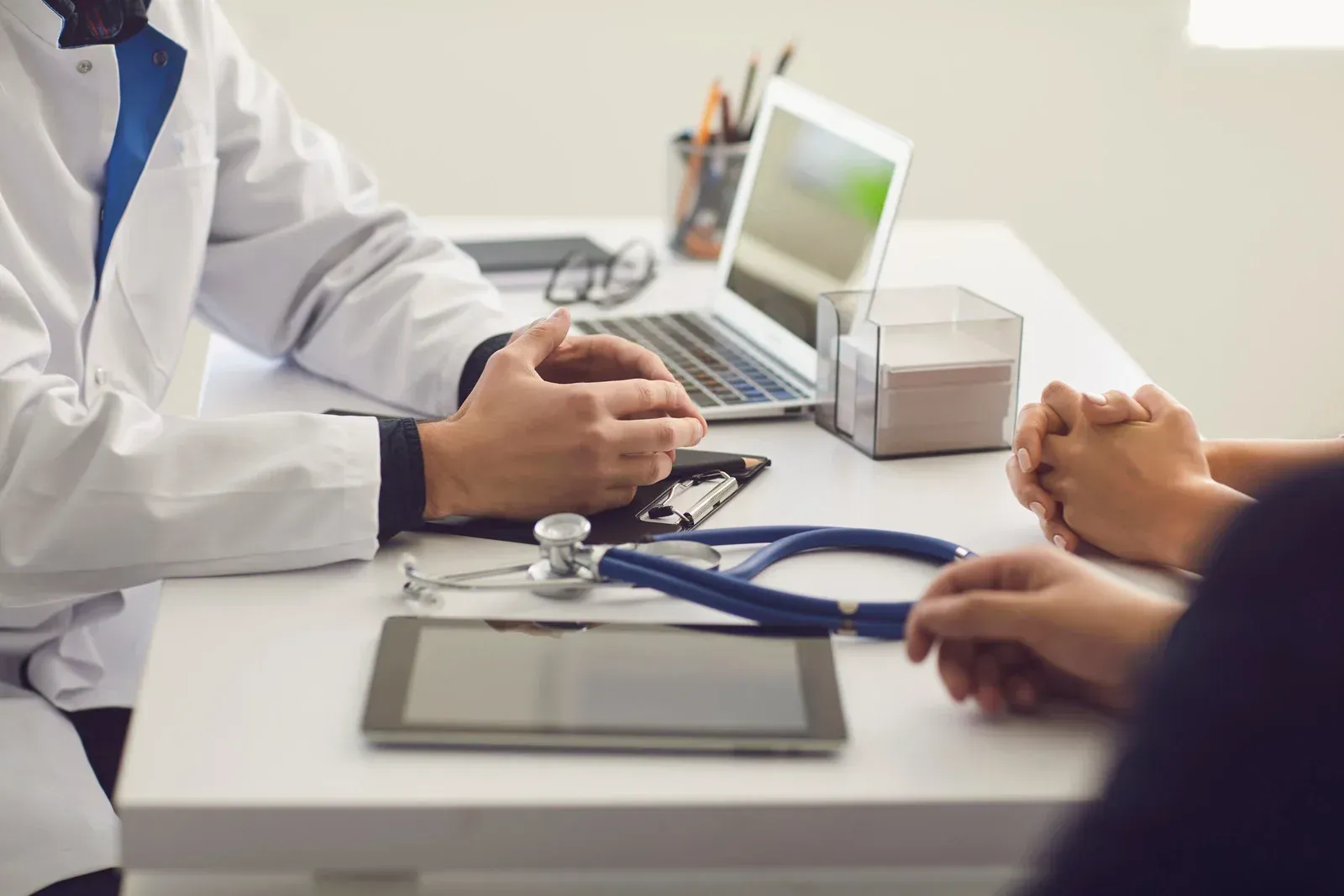 Doctor and patient in a consultation. Doctor in white coat gestures, patient's hands clasped. Medical tools on table.
