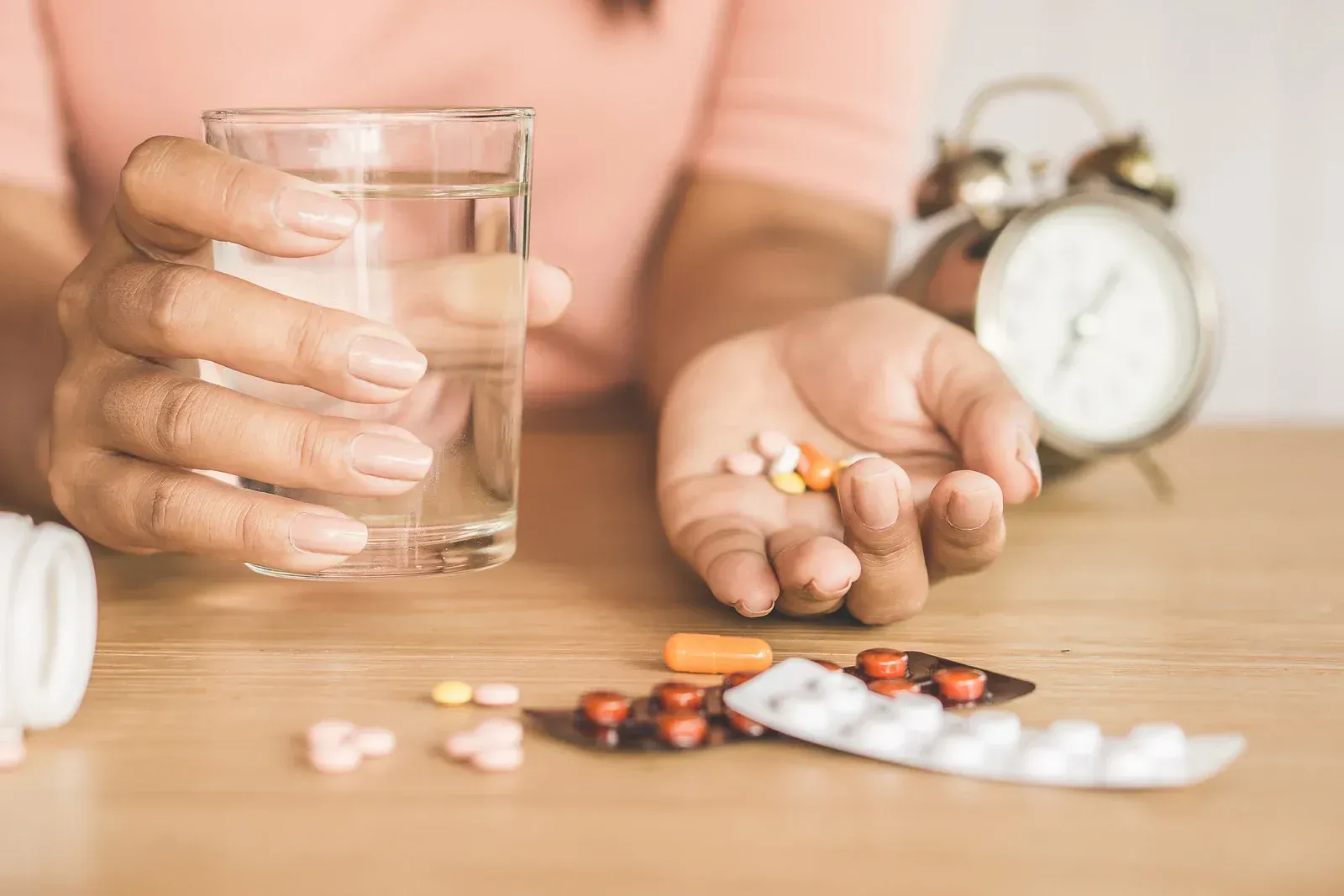 Person holding pills in one hand and glass of water in the other, with alarm clock and medicine on table.