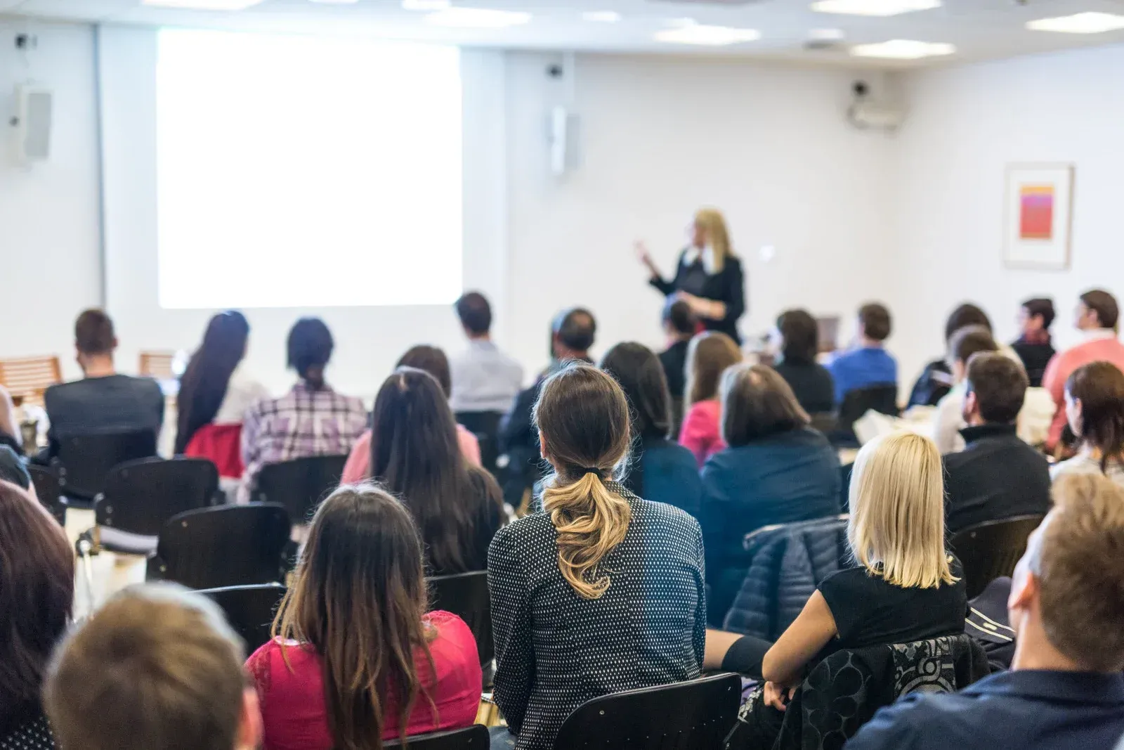 Audience seated, facing a woman presenting at a conference. Blank projector screen.