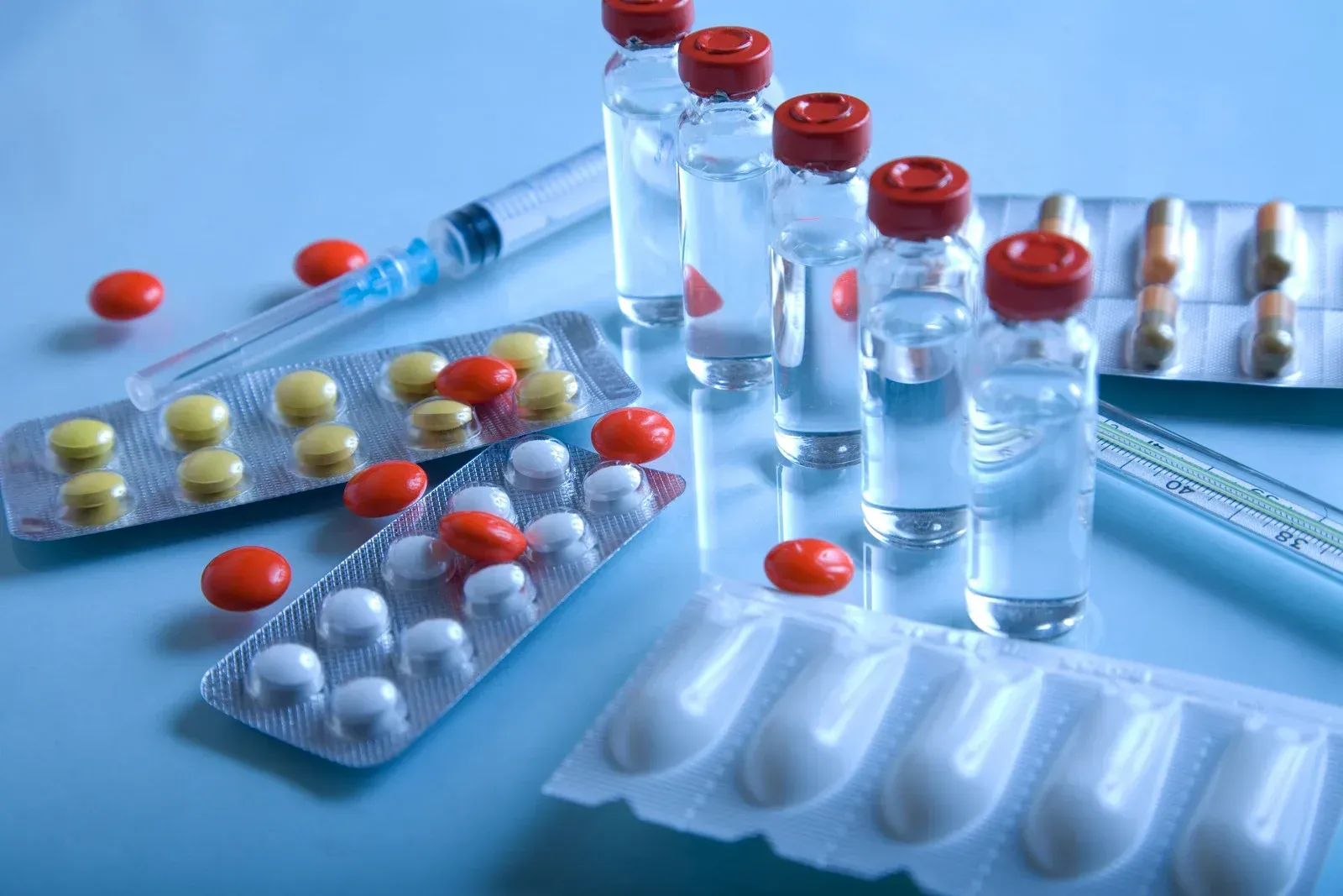 Pills, vials of liquid medicine, a syringe, and suppositories on a light blue surface.