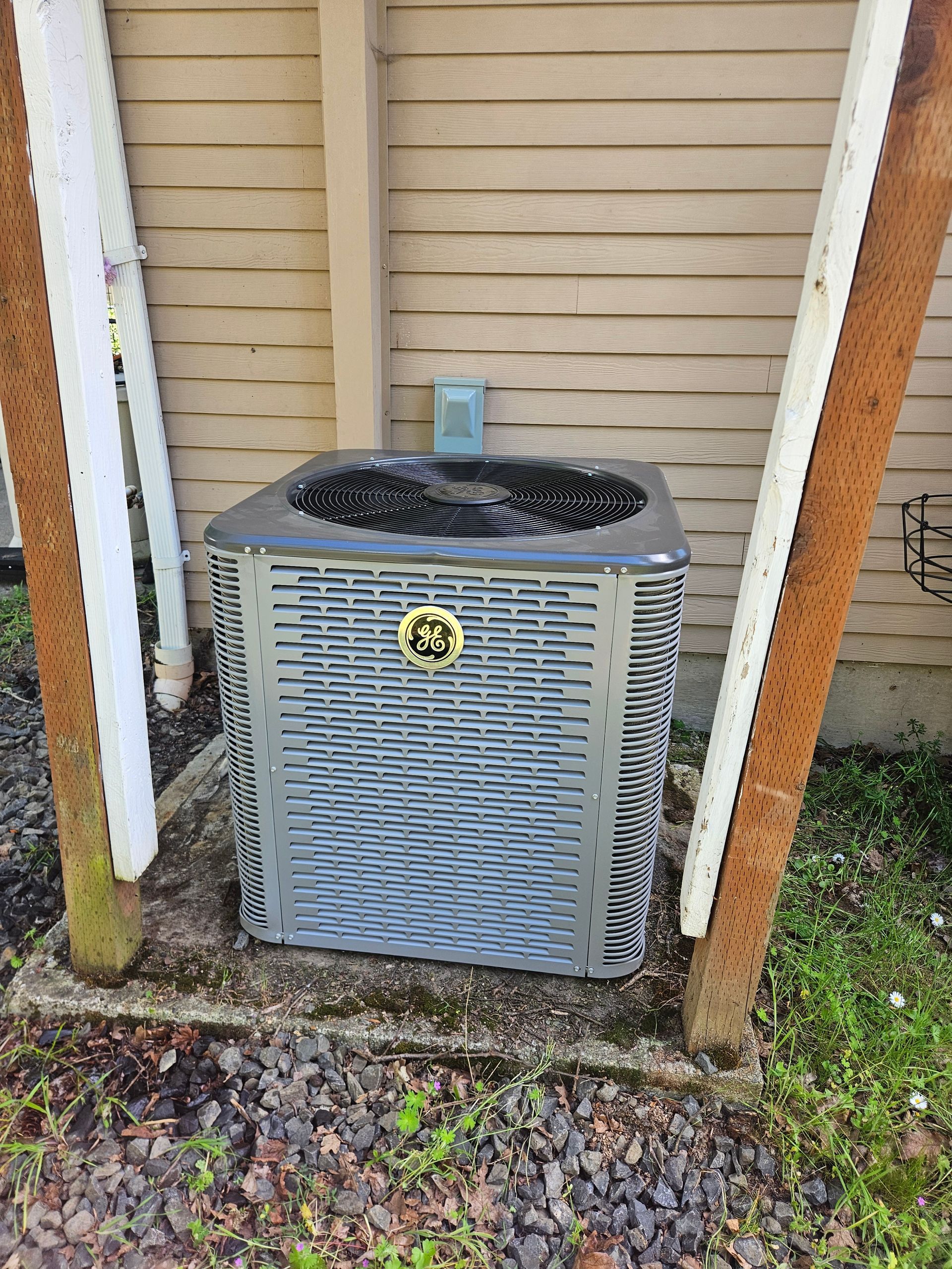 Gray air conditioning unit on concrete base, between wooden posts, near a building.