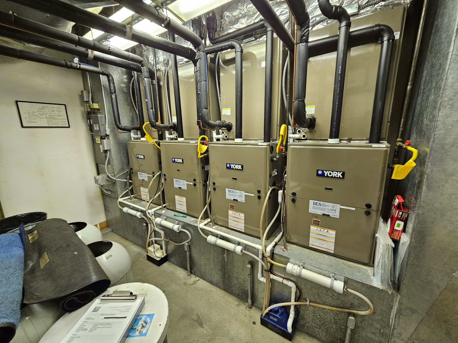 Four furnaces installed in a utility room. Black pipes and white PVC tubing are visible.