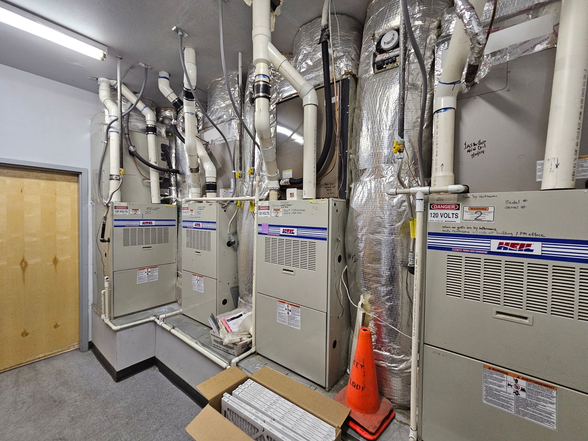 Furnaces in a utility room with vents and piping. A closed wooden door is on the left.