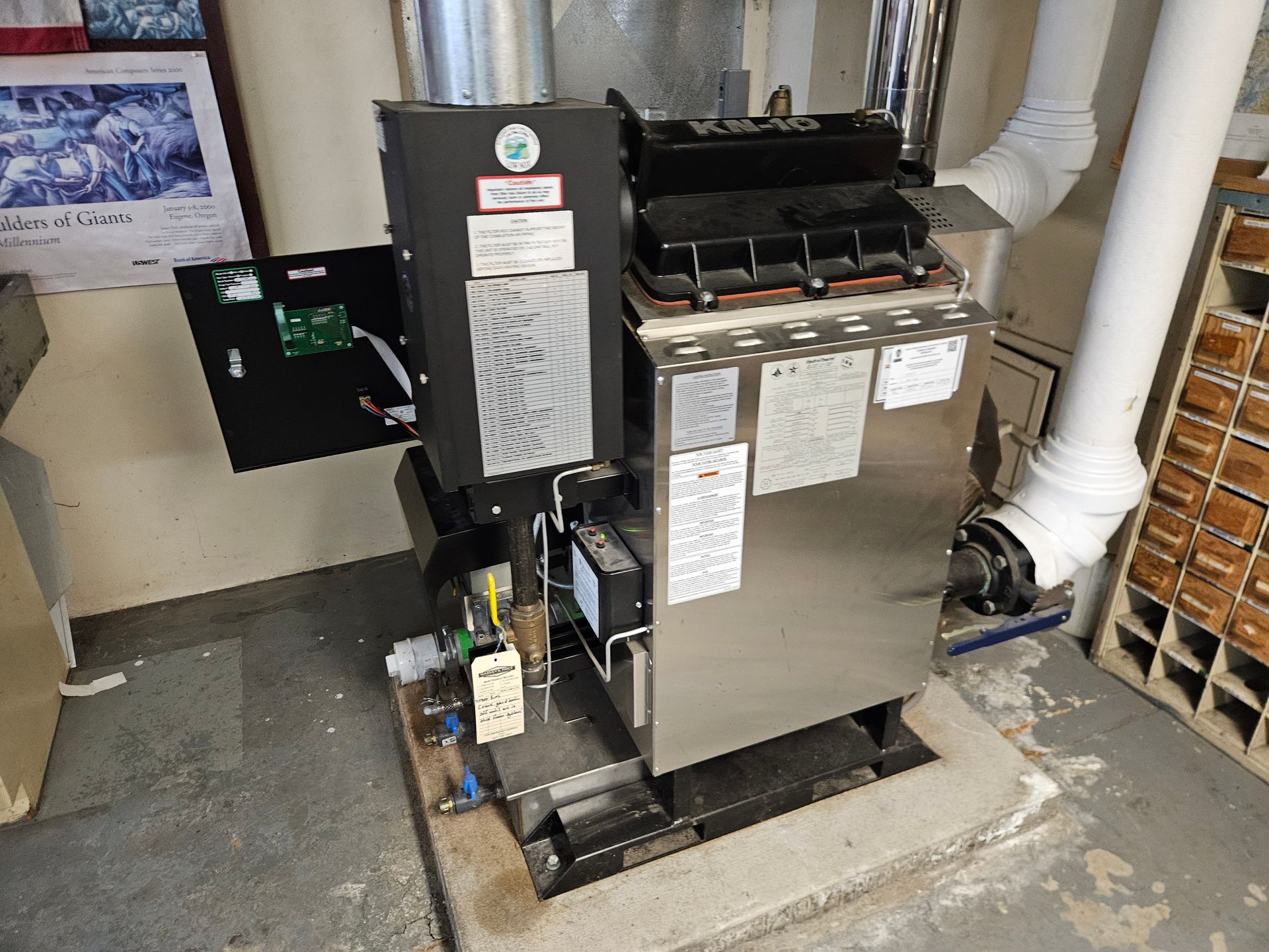 A wood pellet boiler, with open control panel, silver body, and white exhaust pipe, in a utility room.
